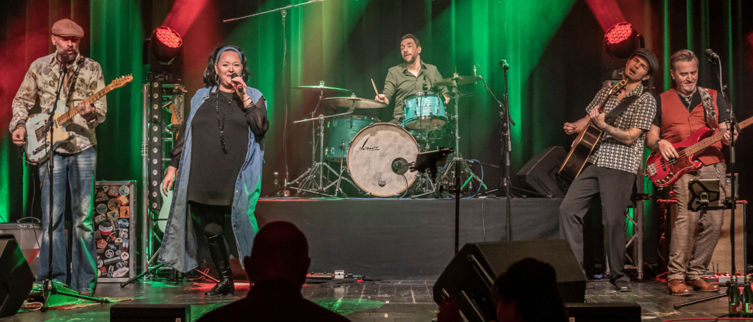 Eine Sängerin steht mit einer Band auf der Bühne und tritt unter grünen und roten Lichtern auf. Ein Schlagzeuger und ein Gitarrist begleiten sie auf der Bühne.