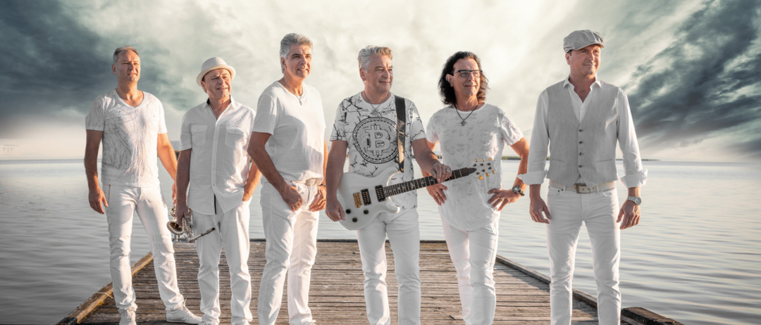 Eine Gruppe von Männern in weißen Kleidern steht auf einem Steg am Wasser, einer hält eine Gitarre, ein anderer eine Trompete.