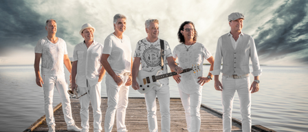 Eine Gruppe von Männern in weißen Kleidern steht auf einem Steg am Wasser, einer hält eine Gitarre, ein anderer eine Trompete.