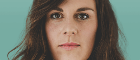 Eine Frau mit braunen, welligen Haaren und einem weißen Hemd schaut direkt in die Kamera. Sie hat rosa Lippen und braune Augen.