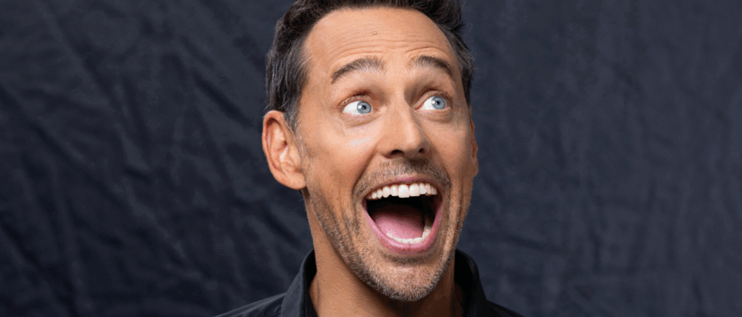 Ein Mann mit kurzen schwarzen Haaren, einem Bart und blauen Augen lacht mit offenem Mund und schaut nach oben. Er trägt ein schwarzes Polo-Shirt.