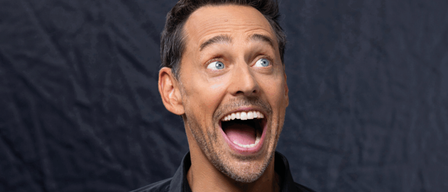 Ein Mann mit kurzen schwarzen Haaren, einem Bart und blauen Augen lacht mit offenem Mund und schaut nach oben. Er trägt ein schwarzes Polo-Shirt.