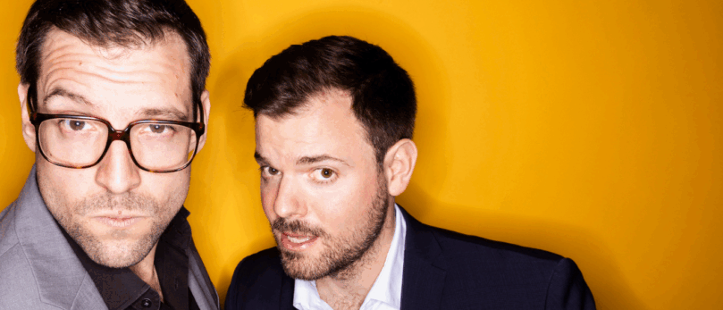 Zwei Männer stehen Seite an Seite vor einem leuchtend gelben Hintergrund. Einer trägt einen grauen Anzug und eine Brille, der andere einen dunklen Anzug mit einem weißen Hemd. Beide schauen direkt in die Kamera.