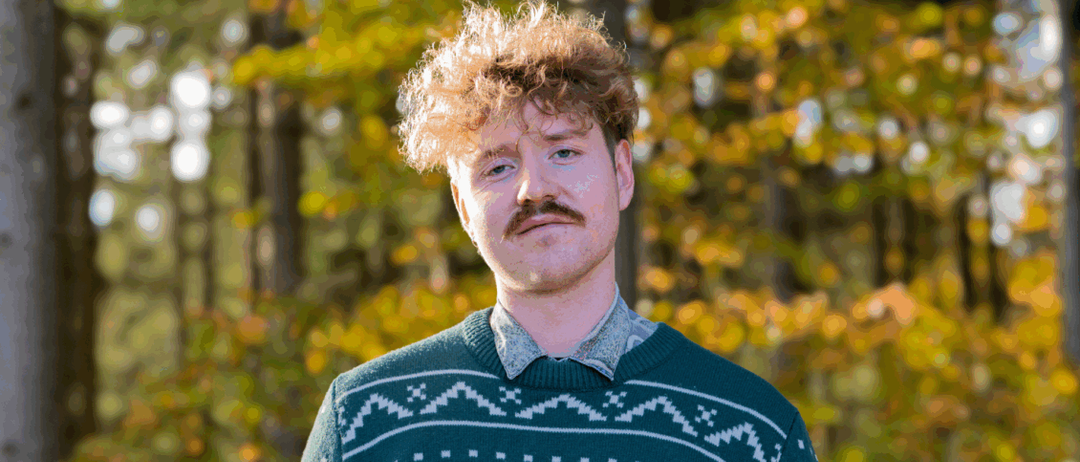 Ein Mann mit lockigem Haar und einem Schnurrbart steht draußen vor einem Hintergrund von Herbstbäumen. Er trägt einen grünen Pullover mit Weihnachtsbaummuster.