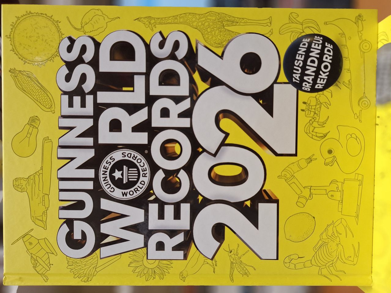 Der Buchdeckel ist gelb mit großen schwarzen Texten, die 'Guinness World Records 2022' lesen. Der Cover enthält verschiedene Illustrationen, darunter einen Skorpion, einen Roboter und eine Zitrone. Das Logo von Guinness World Records befindet sich in der Mitte.