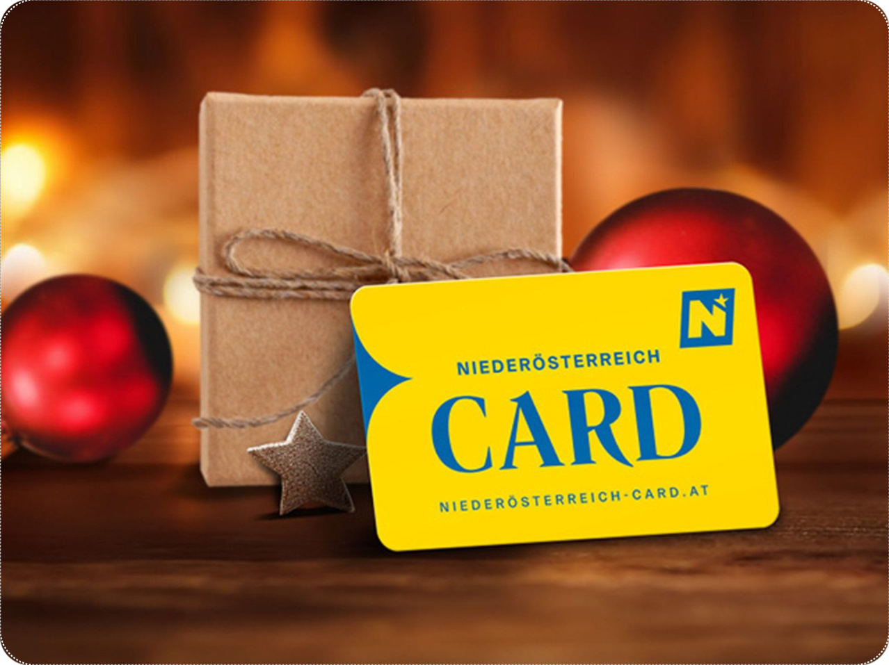 Eine gelbe Niederösterreich Card liegt neben einem verpackten Geschenk und roten Weihnachtskugeln auf einem Tisch.