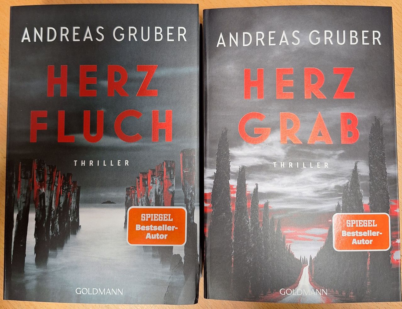 Two book covers titled 'Herz' and 'Herz Grab' by Andreas Gruber, featuring a grey and orange color scheme with landscapes in the background, placed on a wooden surface.