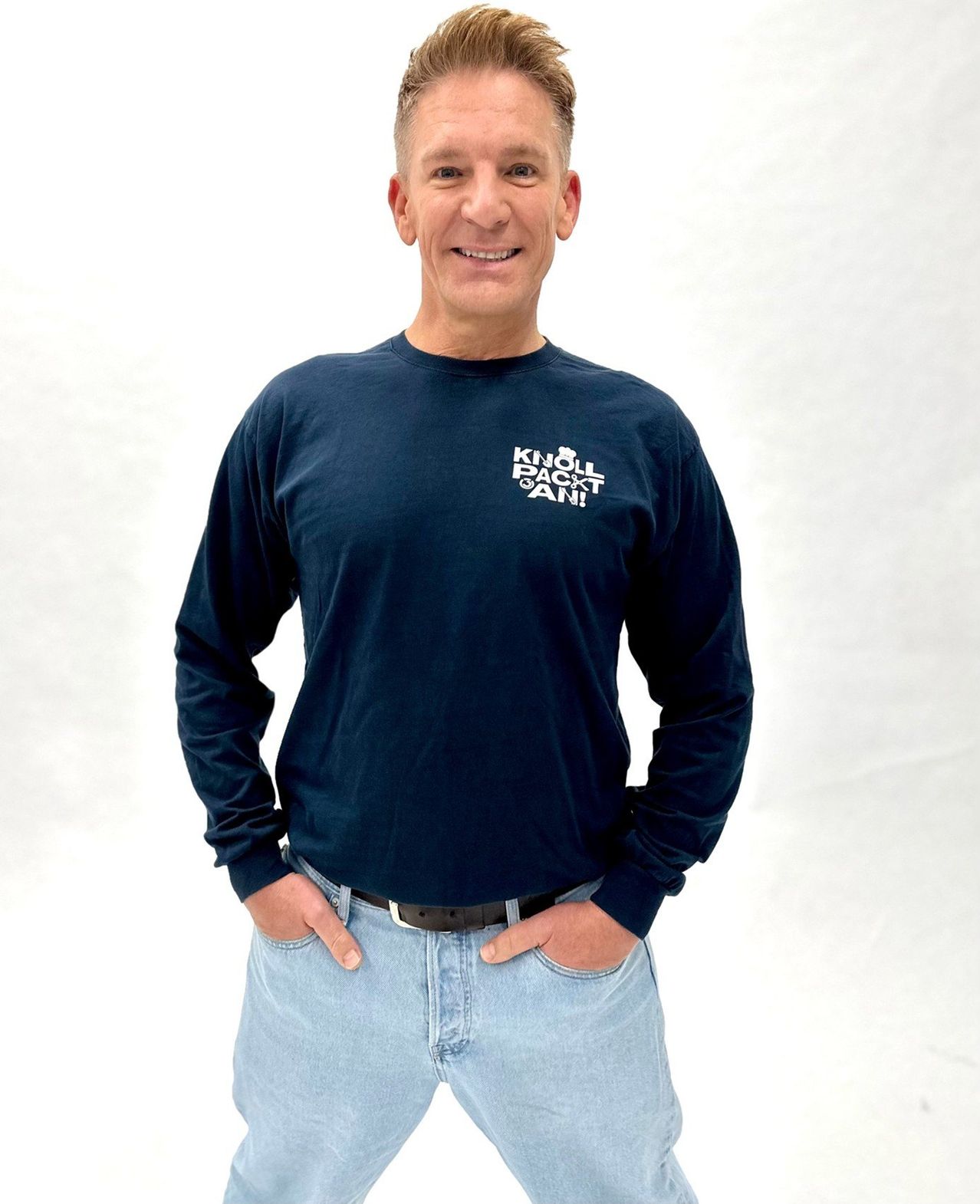 Ein Mann in einem blauen Langarmshirt mit weißem Text 'KNOLL BACKTAN'. Er steht lächelnd mit den Händen in den Taschen vor einem schlichten weißen Hintergrund.