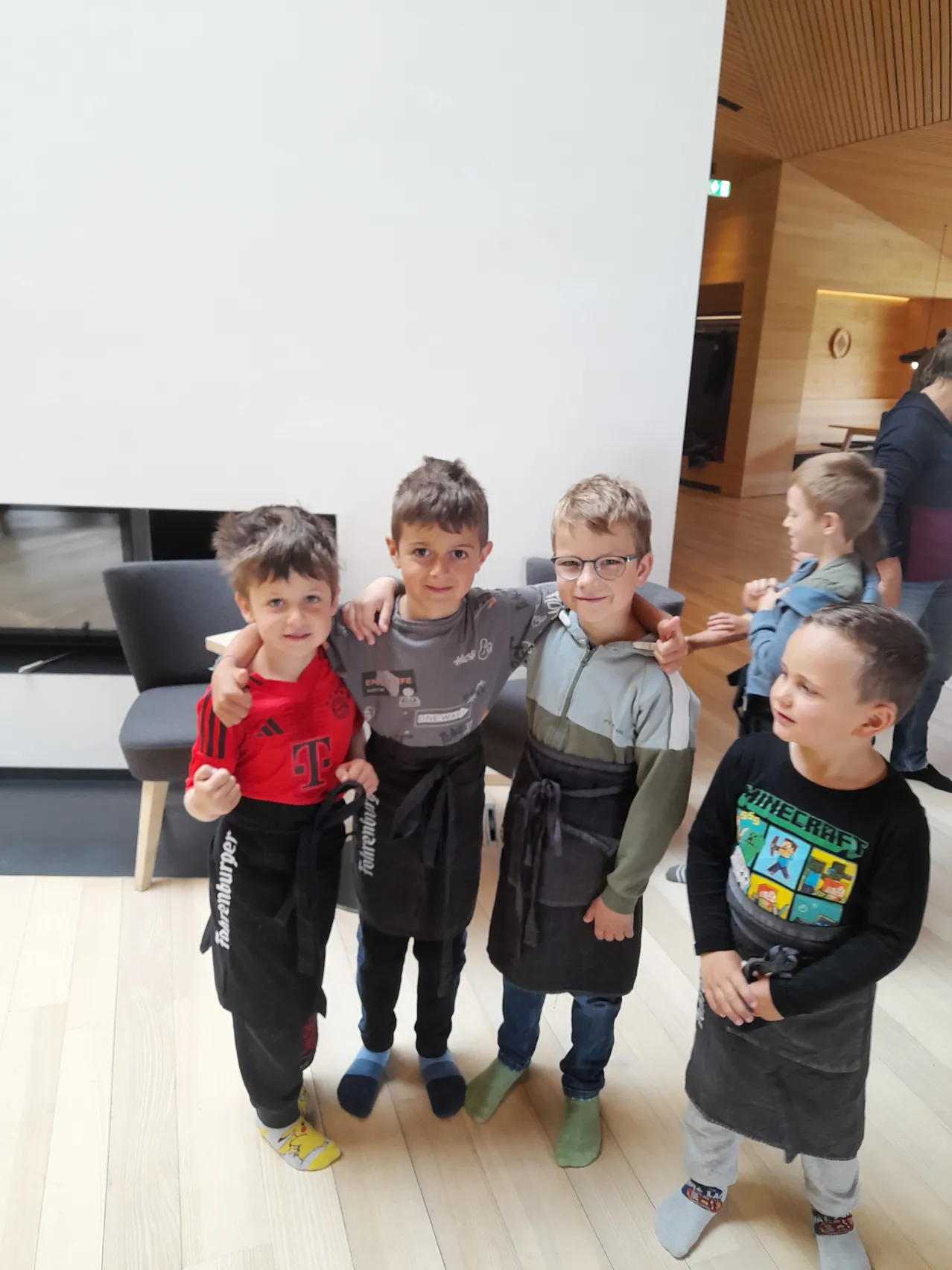Fünf Jungen stehen mit Schürze in einem Raum, einer trägt eine Brille. Hinter ihnen steht eine Person in der Nähe eines Kamins, und eine andere sitzt auf einem Stuhl.