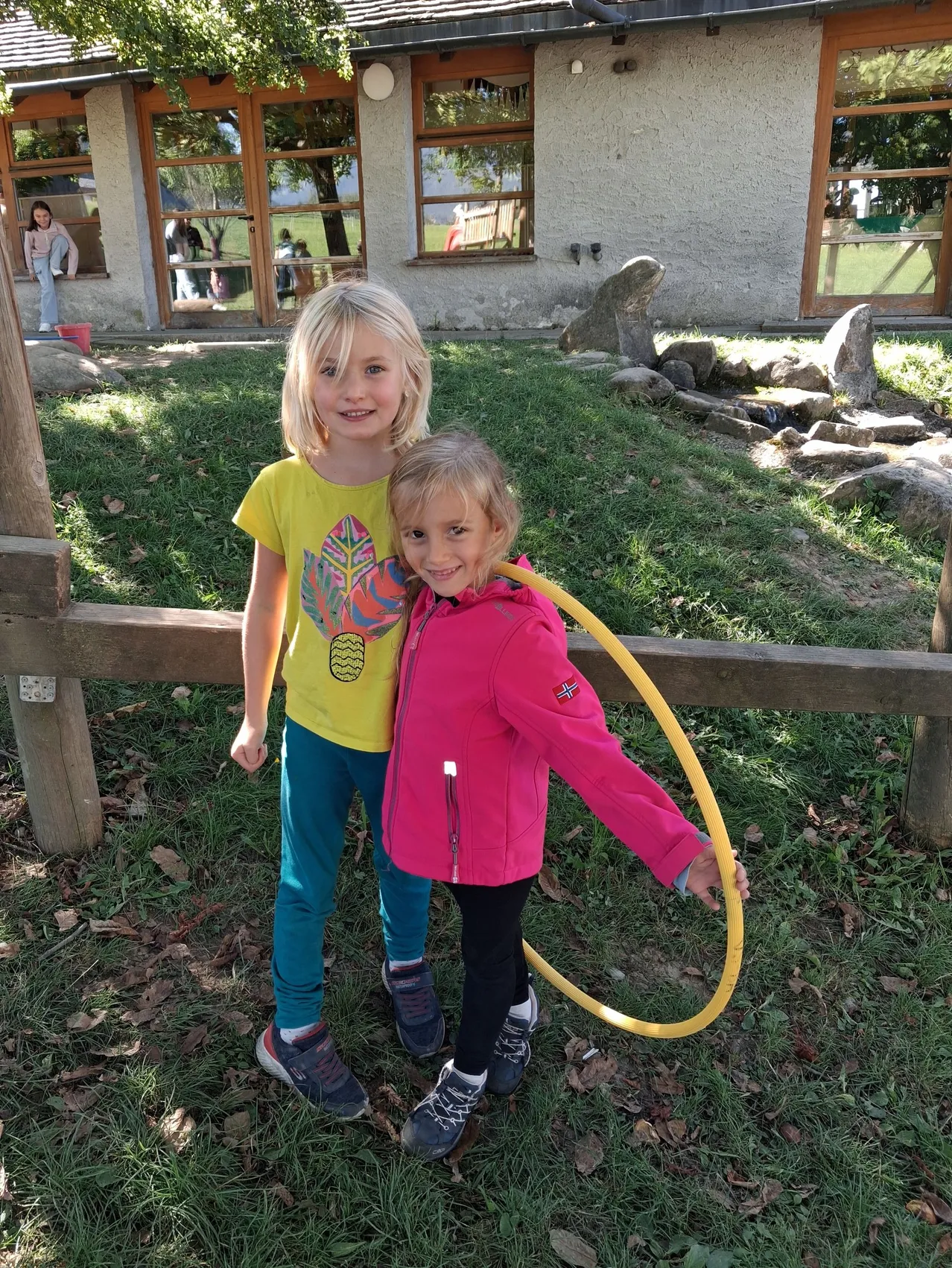 Zwei kleine Mädchen, eines in einem gelben Shirt und das andere in einer rosa Jacke, stehen zusammen in einem Grasbereich mit einem Holzzaun und einem Hula-Hoop.