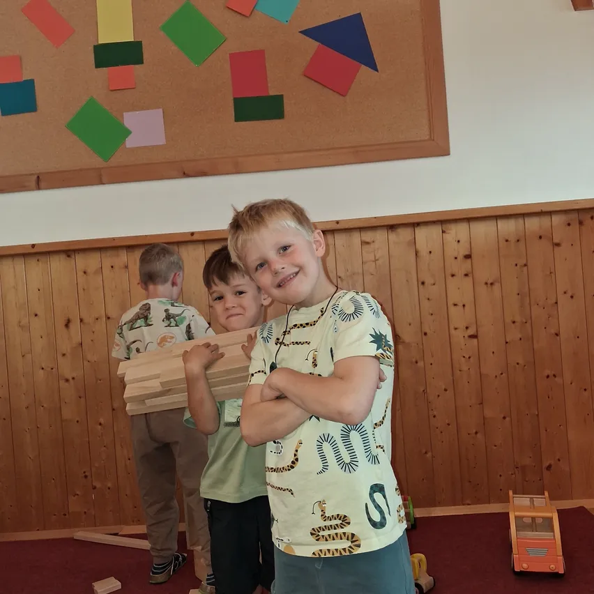 Drei junge Jungen stehen in einem Raum mit Holzwänden, einer hält einen Stapel Holzklötze. Dahinter befindet sich eine Pinnwand mit farbigen Papierformen.