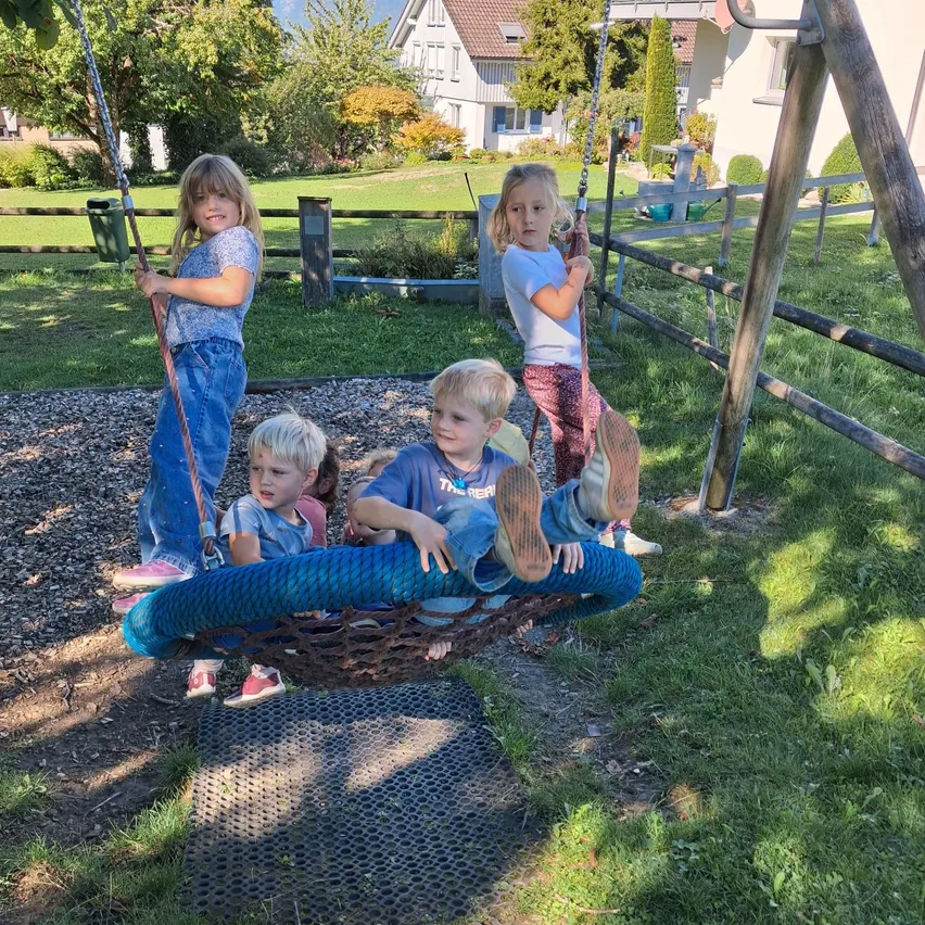 Vier Kinder spielen auf Schaukeln im Garten. Zwei sitzen auf einer Reifenschaukel, eines auf einer normalen Schaukel und eines steht. Dahinter befindet sich ein Holzzaun und ein Haus.