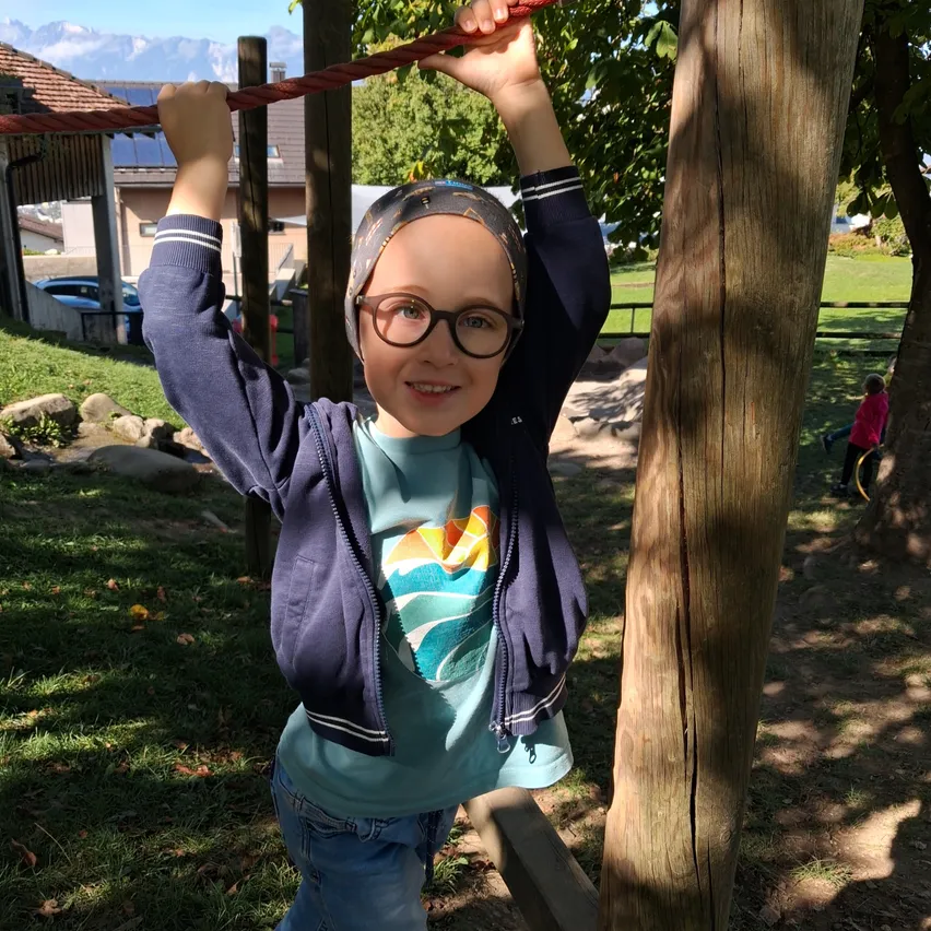 Ein junges Mädchen mit Brille und Kopftuch hängt an einem roten Seil in einem Park. Sie lächelt fröhlich, umgeben von Bäumen und Gras.