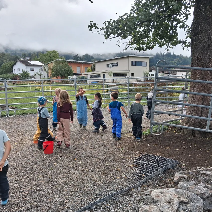 Eine Gruppe von Kindern in einem eingezäunten Bereich, einige in Latzhosen, gehen herum und spielen mit einem roten Eimer.