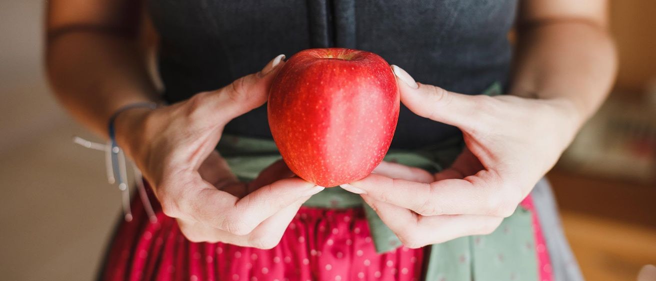 Eine Frau in traditioneller bayerischer Kleidung hält einen roten Apfel und trägt eine Libellenbrosche an ihrer Weste.
