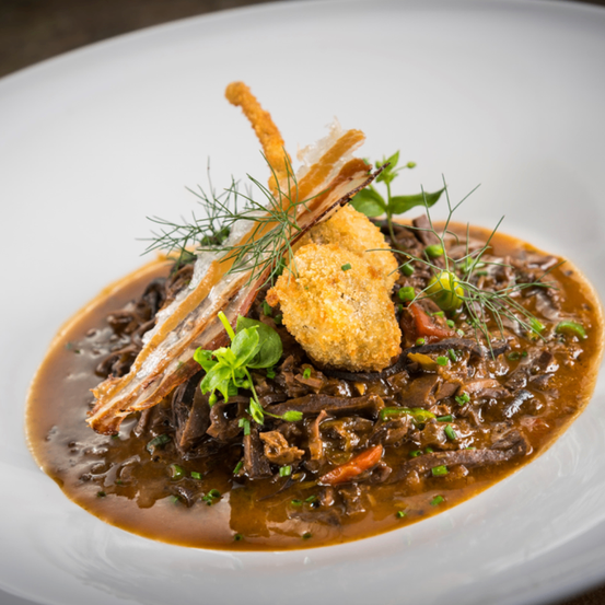 Ein Gourmetgericht mit einer reichhaltigen braunen Sauce, Fleischstücken, knusprigen frittierten Stücken und Kräutern auf einem weißen Teller.