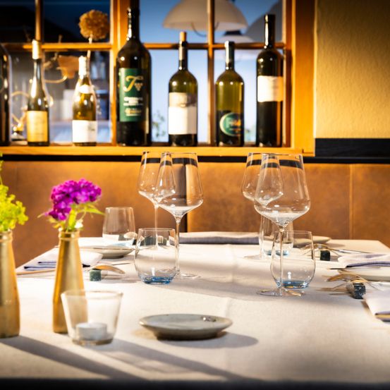 Ein Tisch für eine Mahlzeit mit Weingläsern, Tellern und einer Vase mit lila Blumen. Hinter dem Tisch befindet sich ein Regal mit mehreren Weinflaschen.