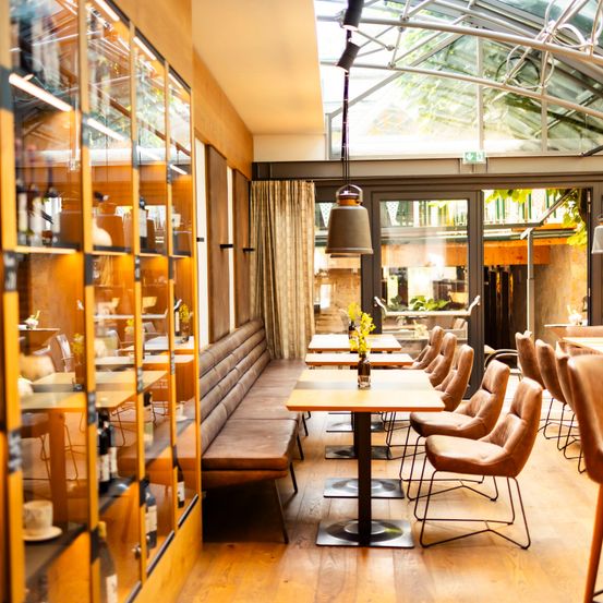 Ein Innenrestaurant mit Holzböden, gläsernen Regalen und einem Glasdach. Der Raum ist mit Tischen, Stühlen und einer langen Bank gefüllt. Der Raum hat eine warme und gemütliche Atmosphäre.