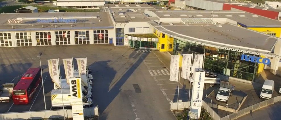 Luftaufnahme eines Industriegebäudes mit vielen Fenstern, einem roten Bus und geparkten Autos. Banner und Fahnen stehen vor dem Gebäude.