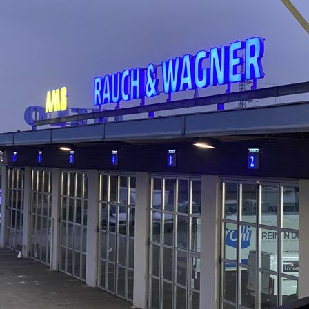 Die Vorderseite eines Gebäudes mit einem blauen Schild, auf dem Rauch & Wagner steht. Die Zahlen 1 bis 4 sind über dem Eingang, und Nummer 2 ist darunter. Das Gebäude hat Glaswände und -türen.