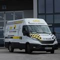 Ein weißer und schwarzer IVECO-Transporter mit kariertem Muster und 'BAUCH & WAGNER' auf der Seite steht vor einem Gebäude mit Glaswänden.