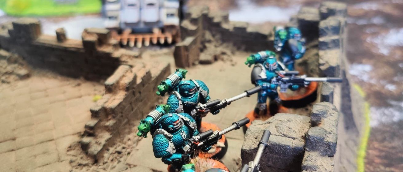 Eine Miniatur-Schlachtfeldszene zeigt Soldaten in blauer Rüstung mit Waffen, die auf einer Steinmauer stehen. Im Hintergrund befinden sich ein Panzer und eine Burg.