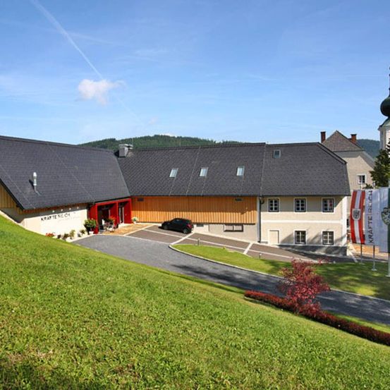 Ein Gebäude mit grauem Dach und rotem Eingang steht auf einem grün bewachsenen Hügel. Ein Auto ist davor geparkt. Ein Turm mit einer Kuppel ist in der Nähe.