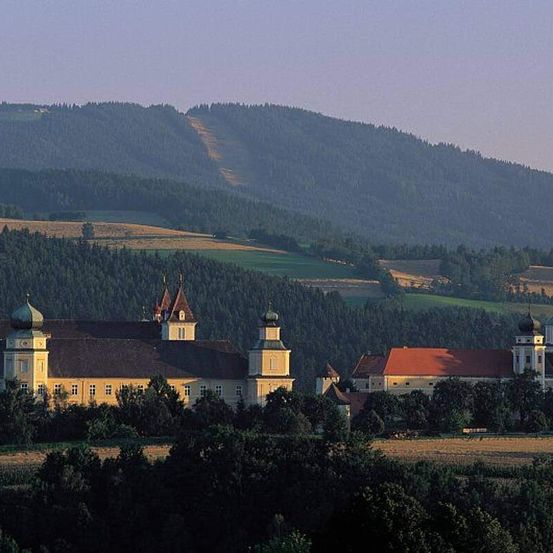 Eine Luftaufnahme eines Klosters, umgeben von Bäumen und Hügeln unter blauem Himmel. Das Kloster hat ein rotes Dach und mehrere Kuppeln.