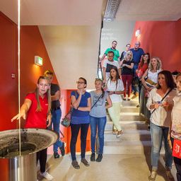 Eine Gruppe von Menschen steht in einem Raum mit einem großen Behälter. Eine Frau in Rot zeigt darauf. Andere schauen die Treppe hinauf, wo ein Mann in Grün nach unten schaut.