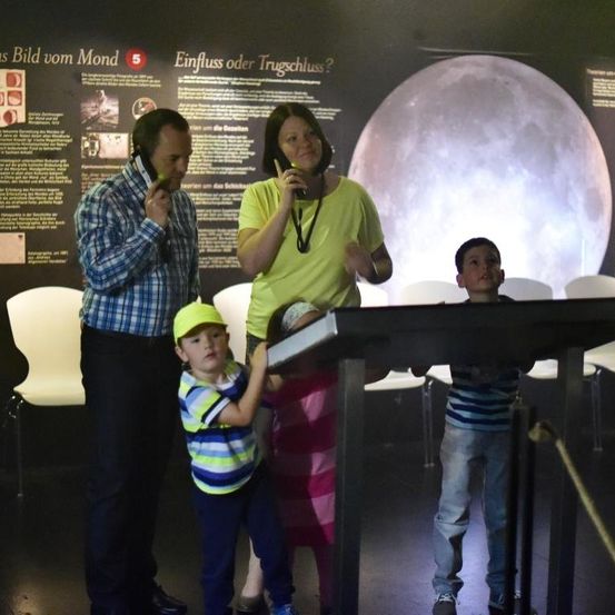 Eine Familie mit vier Personen steht vor einem großen Bildschirm, der den Mond zeigt. Zwei Erwachsene und zwei Kinder, eines benutzt ein Handy und das andere eine Kamera.