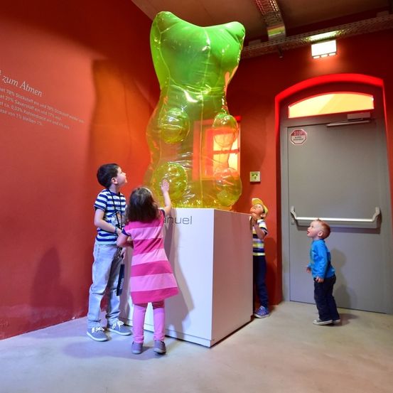 Vier Kinder stehen vor einer großen grünen Gummibär-Skulptur in einem roten Raum. Die Kinder schauen neugierig nach oben zur Skulptur.