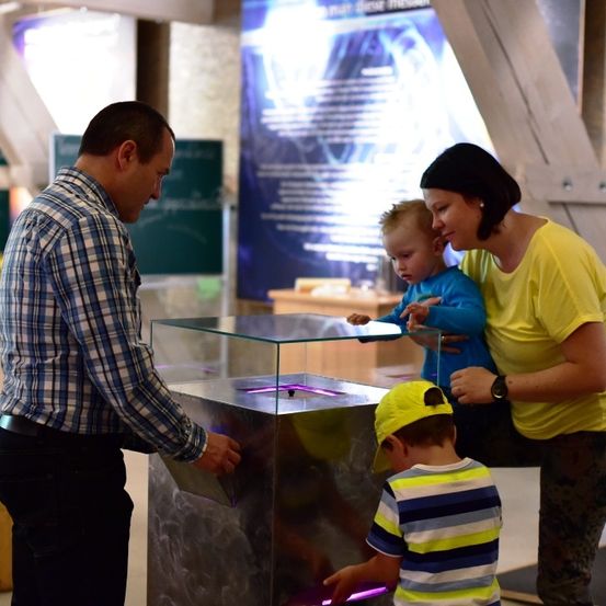 Eine Familie untersucht eine Glasbox mit leuchtendem Licht in einem Museum. Ein Mann, eine Frau und zwei Kinder sind anwesend. Der Mann hält die Box, während die Frau zusieht.