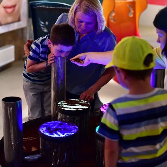 Eine Frau und zwei Kinder interagieren mit einer wissenschaftlichen Ausstellung, der Junge bläst in ein Rohr mit einer leuchtenden Basis.