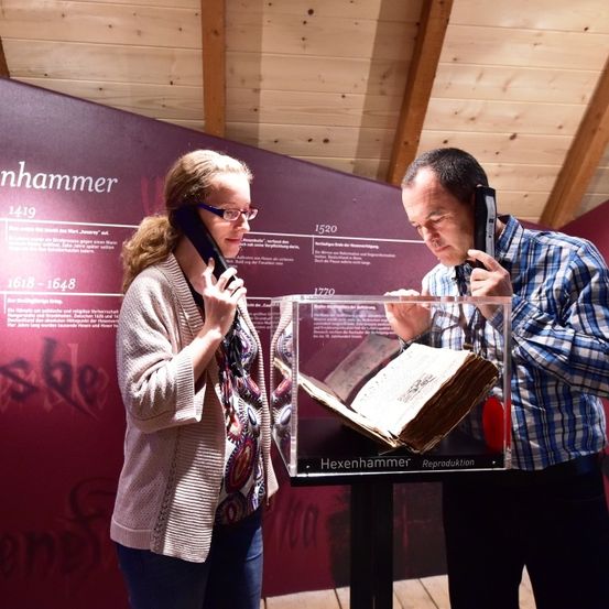 Eine Frau und ein Mann untersuchen ein altes Buch in einer Museumsausstellung, beide halten antike Telefone. Hinter ihnen befindet sich eine Wand mit historischen Daten und Text.