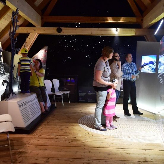 Eine Gruppe von Menschen befindet sich in einem Raum mit Holzboden und -decke. Sie betrachten einen Bildschirm mit Bergblick. Es gibt Stühle, einen Tisch und eine Ausstellung mit Text. Ein Mann hält eine Kamera und eine Frau hält ein Kind.
