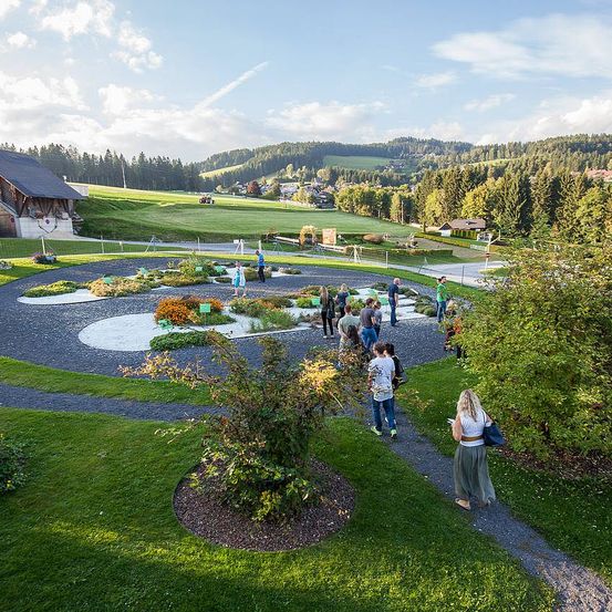 Eine Luftaufnahme eines Gartens mit Menschen, die herumlaufen. Bäume und Pflanzen umgeben den Garten, und ein Gebäude ist in der Ferne zu sehen. Ein großes grünes Feld und Berge sind im Hintergrund sichtbar.