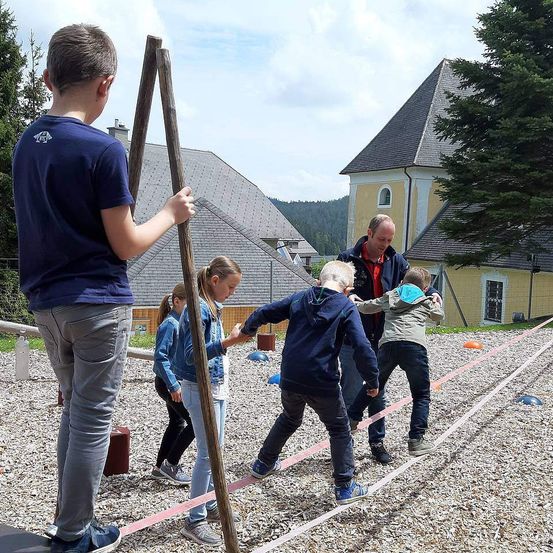 Eine Gruppe von Kindern und Erwachsenen spielt auf einem Kiesfeld mit einem gelben Gebäude im Hintergrund. Die Kinder halten Stöcke und laufen auf einem rosafarbenen Seil.