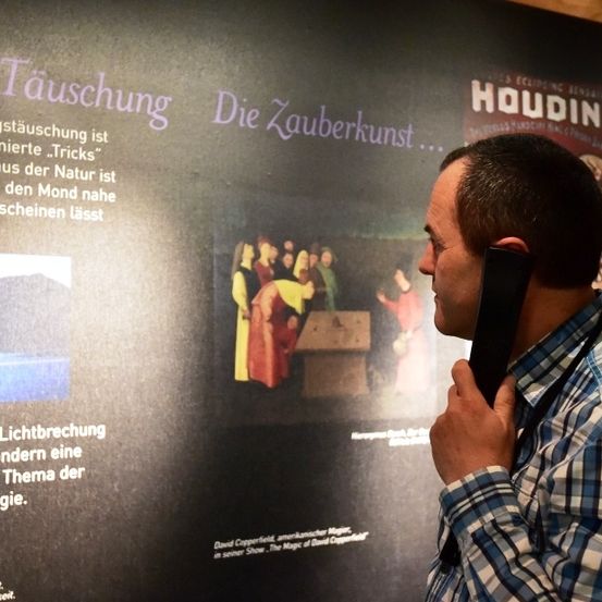 Ein Mann in einem karierten Hemd und mit Kopfhörern steht vor einer Wand mit einer Projektion einer Szene und Text über Lichtbrechung und Psychologie.