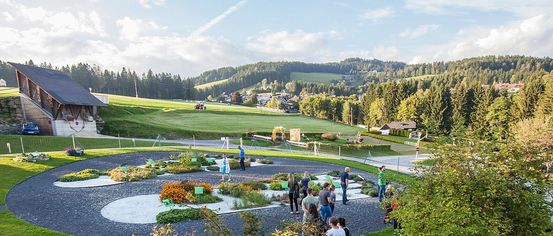 Menschen besuchen einen Garten, umgeben von grüner Wiese und Bäumen. In der Ferne fährt ein Traktor auf einem Feld. Der Himmel ist bewölkt.