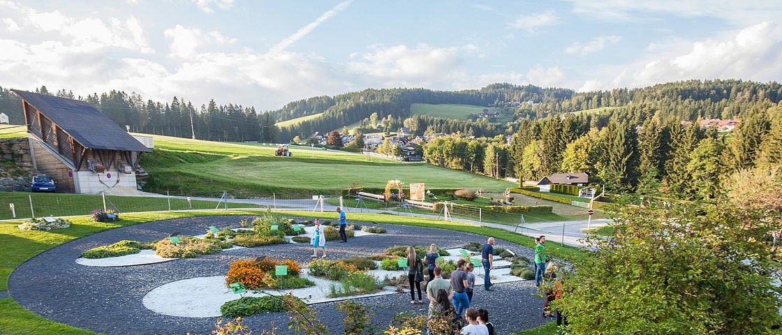 Menschen besuchen einen Garten, umgeben von grüner Wiese und Bäumen. In der Ferne fährt ein Traktor auf einem Feld. Der Himmel ist bewölkt.
