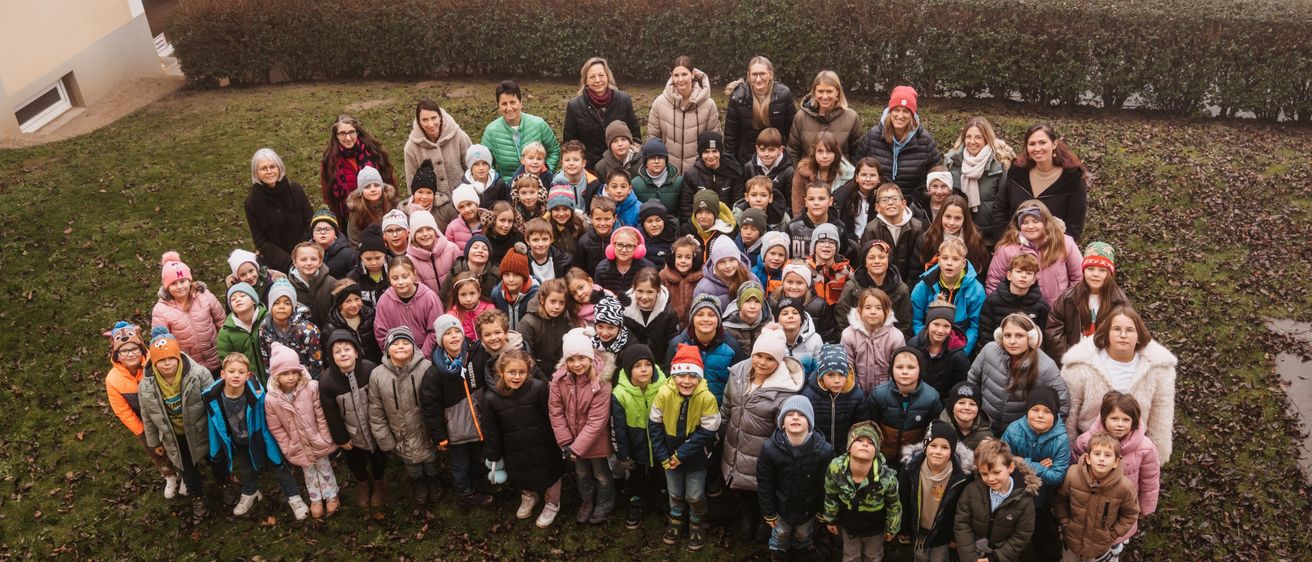 A large group of children and adults are gathered outdoors, posing for a photo. They are dressed in winter clothing, some with hats and scarves. They are standing in front of a building with a green gate.