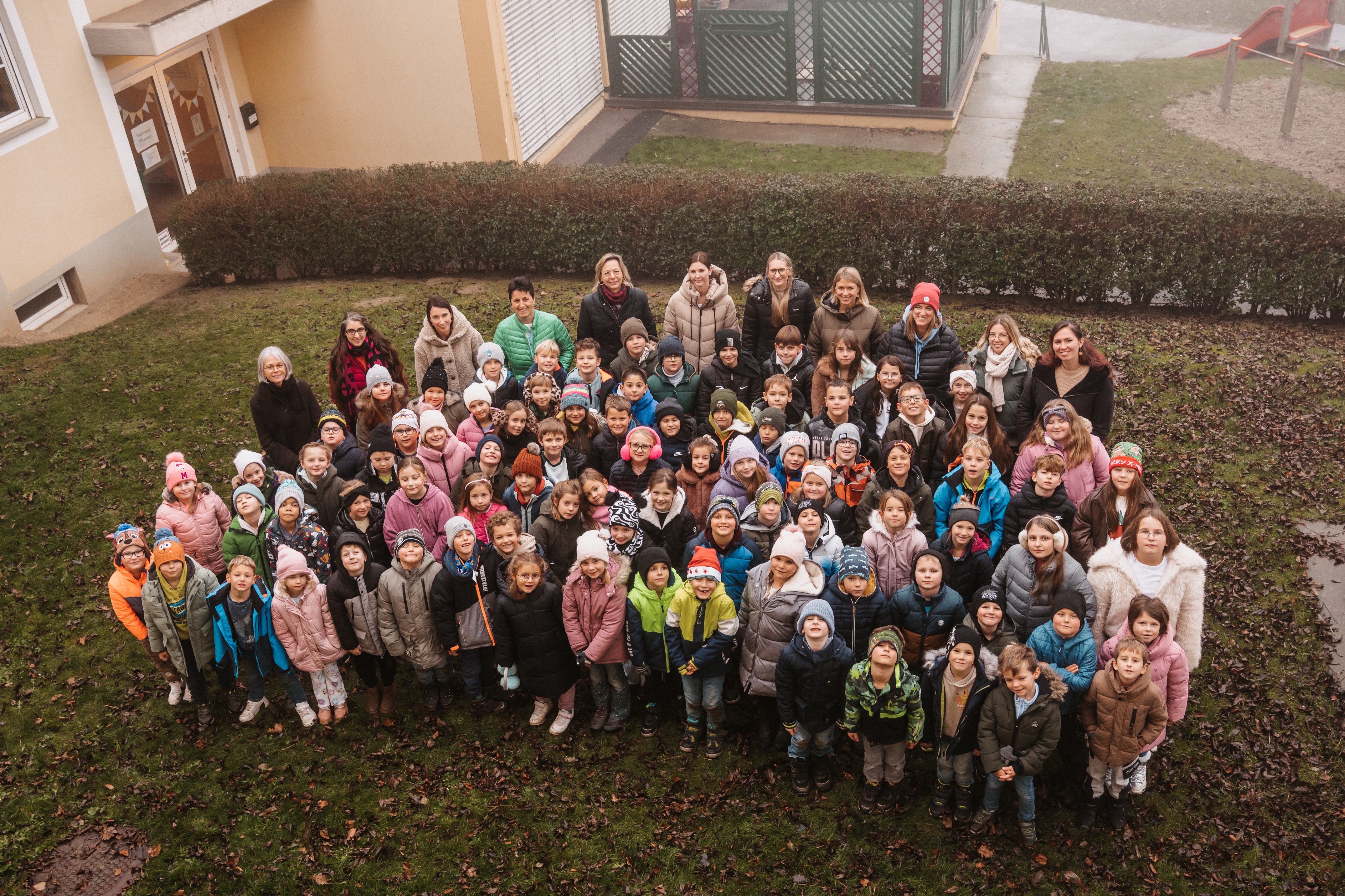 Eine große Gruppe von Kindern und Erwachsenen versammelt sich auf einem Rasenfeld, wahrscheinlich für ein Gruppenfoto. Sie sind in Winterkleidung gekleidet, einige tragen Mützen und Brillen. Dahinter steht ein Gebäude mit einem grünen Tor und Büschen.