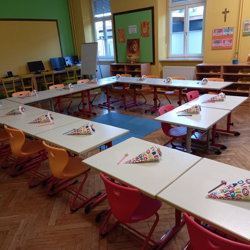 Ein Klassenzimmer mit Tischen und Stühlen in Reihen angeordnet, farbenfrohe Partyhüte auf jedem Schreibtisch, grüne und gelbe Wände, eine Tafel und ein Kreuz an der Wand.
