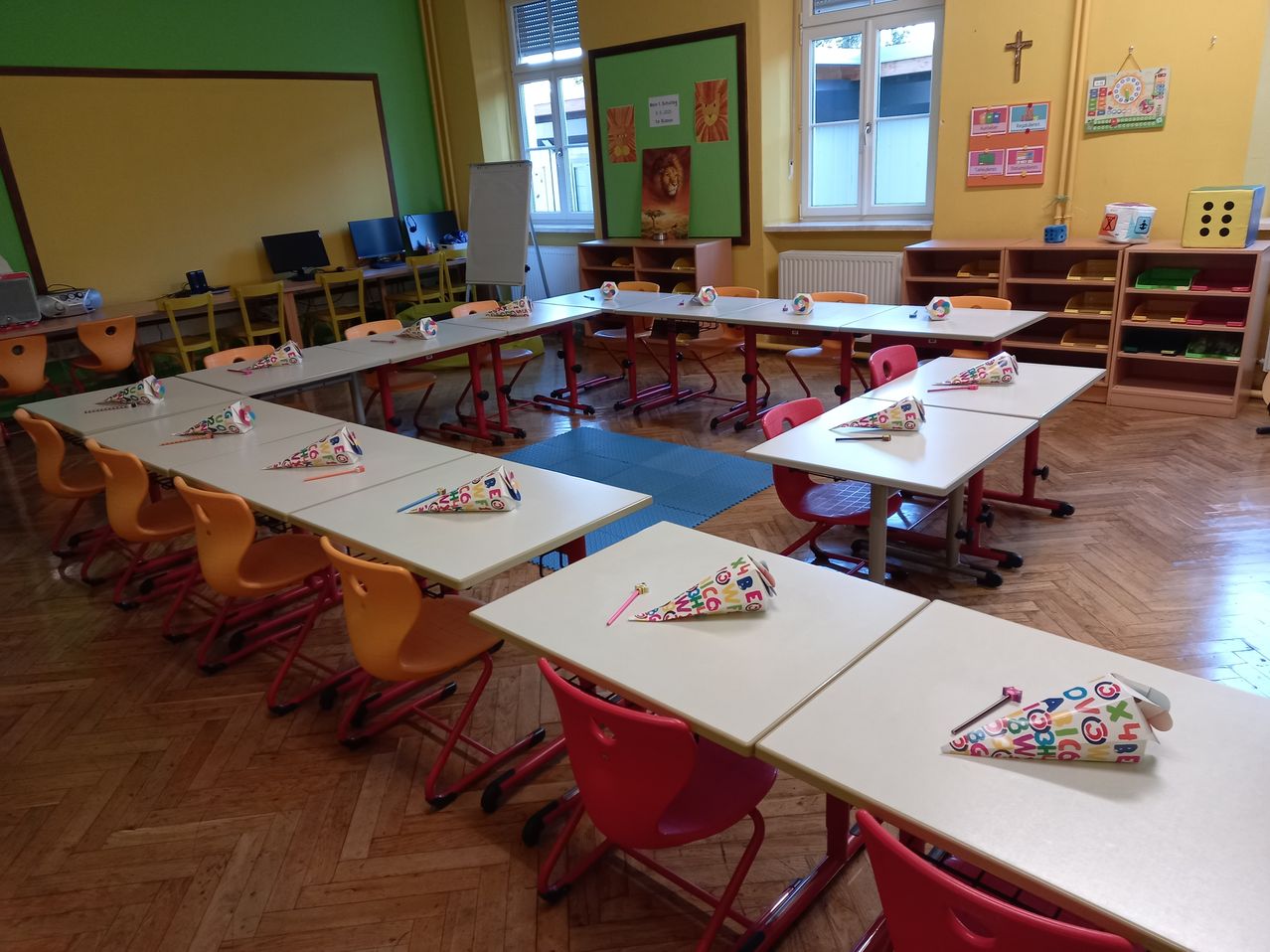 Ein Klassenzimmer mit Tischen und Stühlen in Reihen angeordnet, farbenfrohe Partyhüte auf jedem Schreibtisch, grüne und gelbe Wände, eine Tafel und ein Kreuz an der Wand.