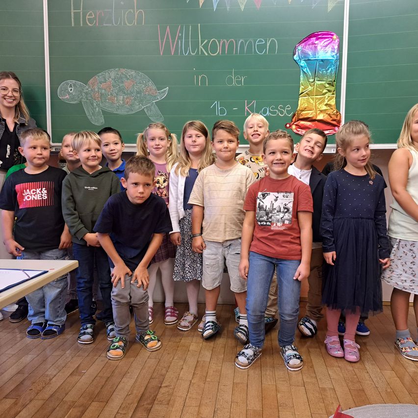 Eine Gruppe von Kindern steht in einem Klassenzimmer und lächelt. Eine Tafel hinter ihnen sagt Herzlich Willkommen in der 1b Klasse. Sie stehen vor einer Schildkrötenzeichnung und einer Nummer eins Luftballon.