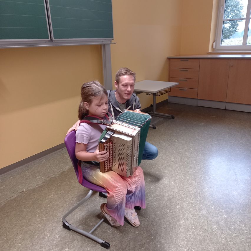 Ein kleines Mädchen sitzt auf einem Stuhl und spielt Akkordeon, während ein Mann ihr von hinten hilft. Sie befinden sich in einem Raum mit gelben Wänden und einem Fenster.