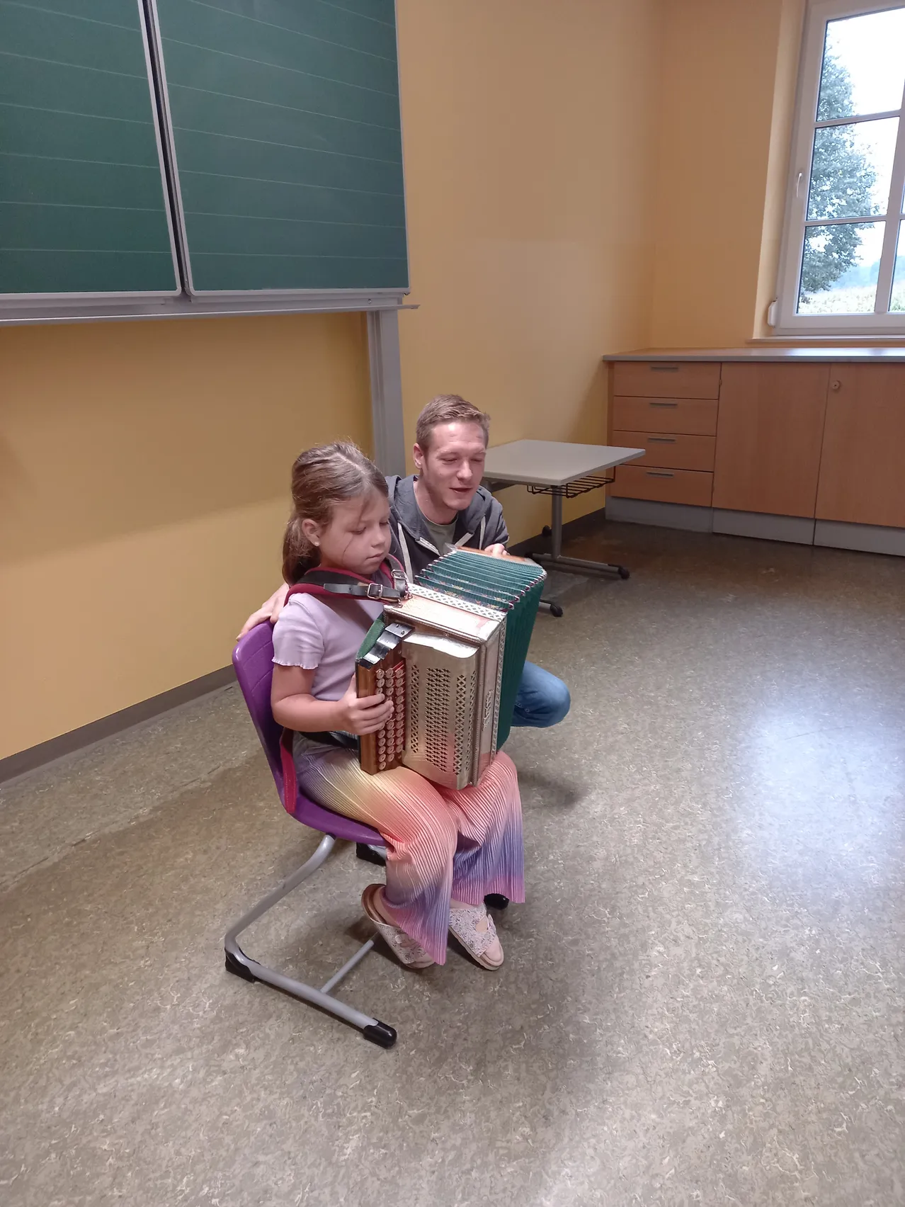 Ein kleines Mädchen sitzt auf einem Stuhl und spielt Akkordeon, während ein Mann ihr von hinten hilft. Sie befinden sich in einem Raum mit gelben Wänden und einem Fenster.