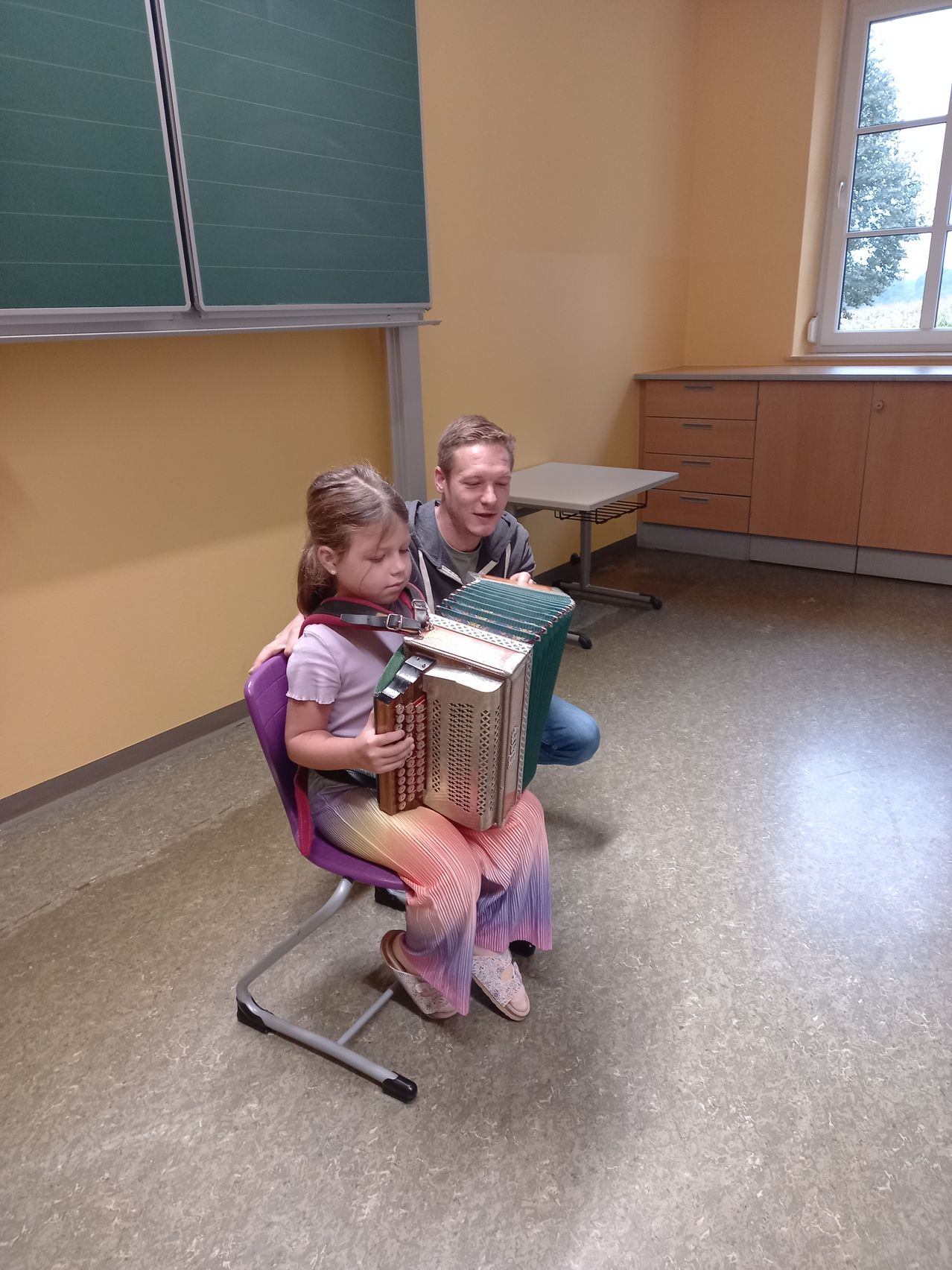 Ein kleines Mädchen sitzt auf einem Stuhl und spielt Akkordeon, während ein Mann ihr von hinten hilft. Sie befinden sich in einem Raum mit gelben Wänden und einem Fenster.