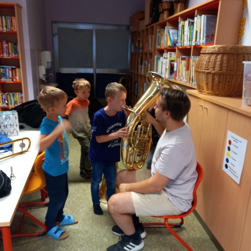 Ein Mann spielt ein Waldhorn, während drei junge Jungen zusehen und zuhören. Sie befinden sich in einem Raum mit Bücherregalen voller Bücher und einem Korb obenauf.