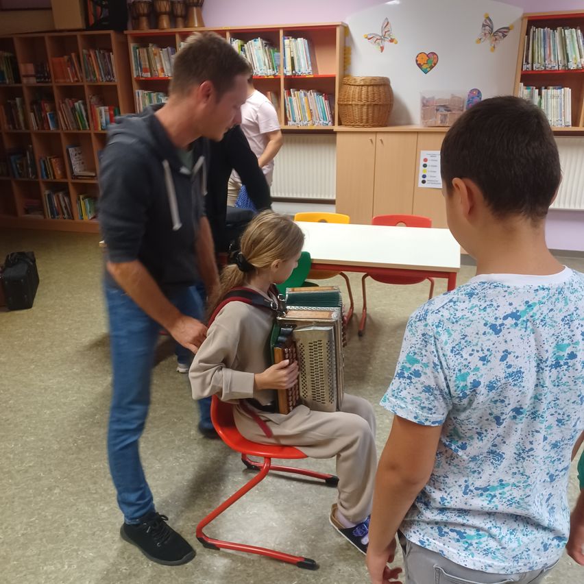 In einer Bibliothek hilft ein Mann einem jungen Mädchen, ein Akkordeon zu spielen, während ein Junge aufmerksam zusieht. Im Hintergrund sind Regale voller Bücher.